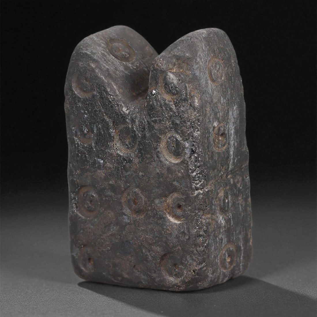 A Near Eastern Jet Idol, 2nd-1st Millennium BCE, 63LD: Carved from dark jet with a matte, weathered surface, the object is shaped as a block-like figure with a bifurcated upper section. The four sides are decorated with regularly spaced circular depressio