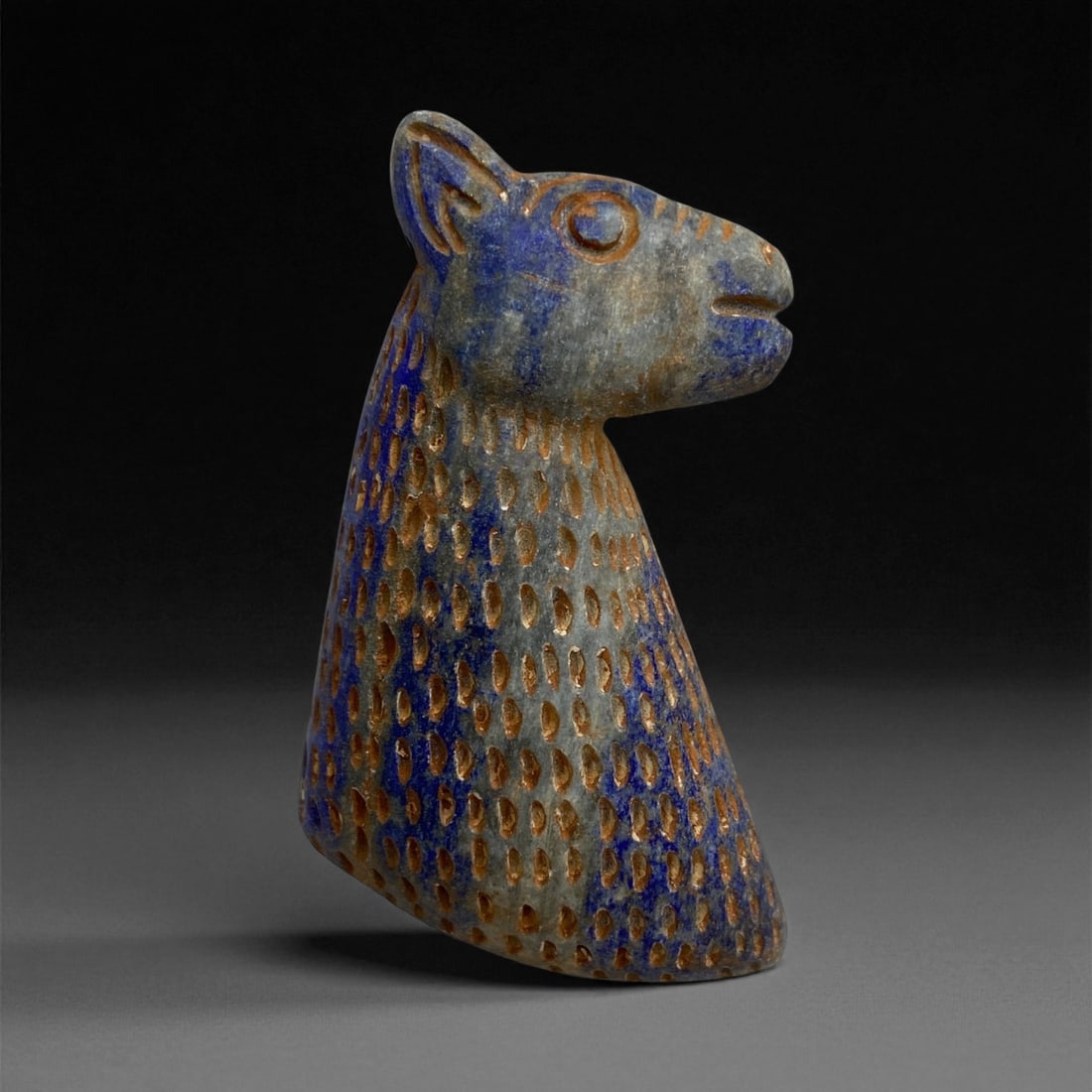 A Near Eastern Lapis Lazuli Feline Plaque, 2nd Millennium BCE, 47g, 27EE: Carved from lapis lazuli with areas of deep blue and lighter veining, the object is shaped as a stylized feline head and elongated tapering body. The surface is covered with evenly spaced ovoid depres