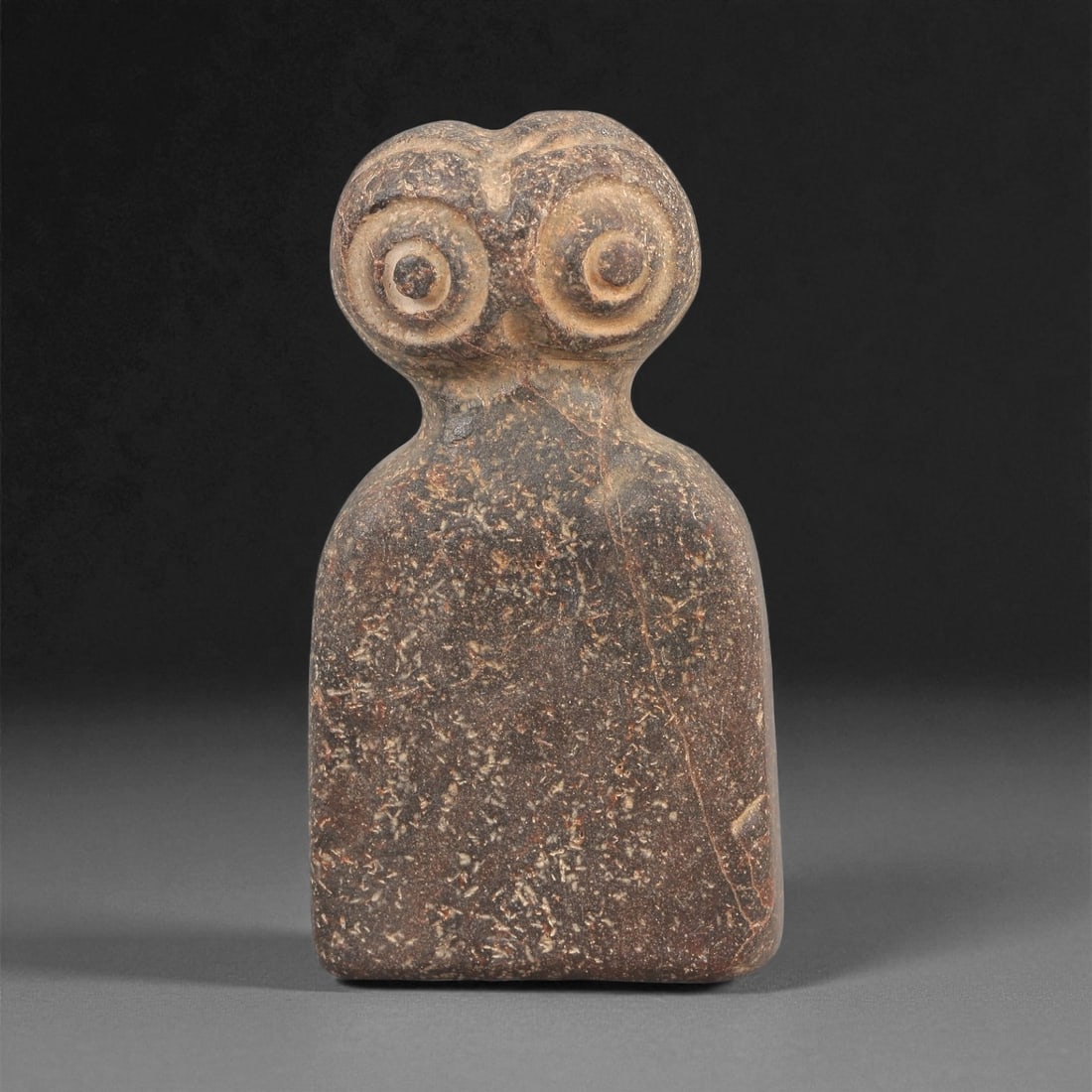 A Near Eastern Stone Eye Idol, 4th-3rd Millennium BCE, 81LD: Carved from a dark brown stone with scattered light inclusions, the object is shaped with a rounded upper section bearing a pair of deeply recessed concentric eyes. The lower body is rectangular with 