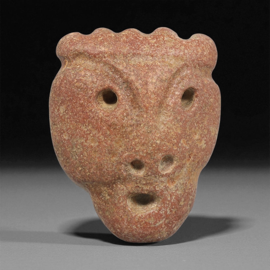 A Mediterranean Stone Maskette, 12th-17th Century, 97LC: Carved from a reddish-brown stone with fine granular texture, the maskette is shaped with a rounded upper outline featuring a scalloped crest. The facial features are executed in shallow relief and dr