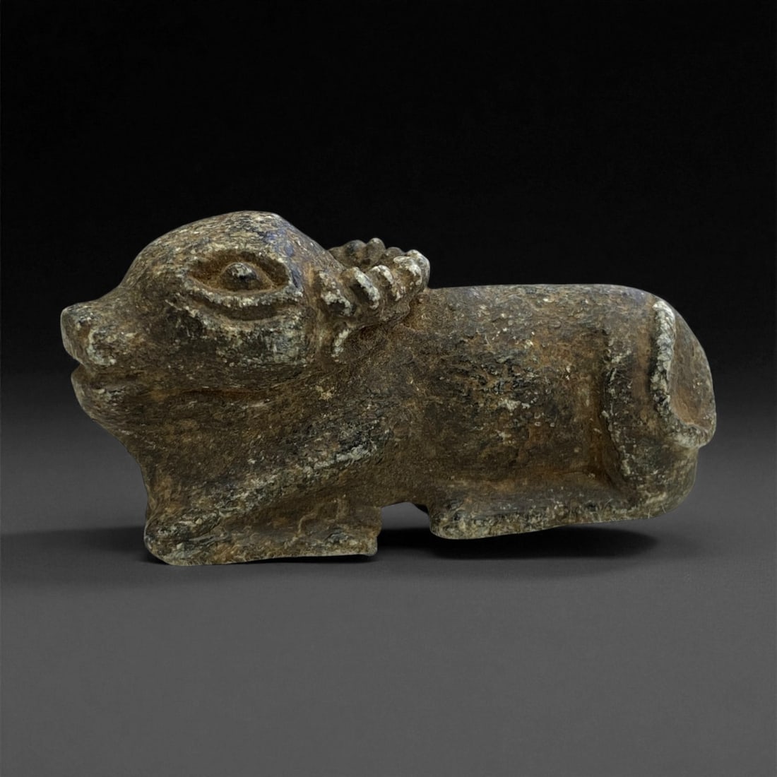 A Near Eastern Stone Buffalo Effigy, 2nd-1st Millennium BCE, 39g, 26EE: Carved from a mottled dark stone with lighter inclusions, the figure represents a recumbent bovine with a rounded body and short projecting legs. The head is rendered with pronounced eyes, a slightly 