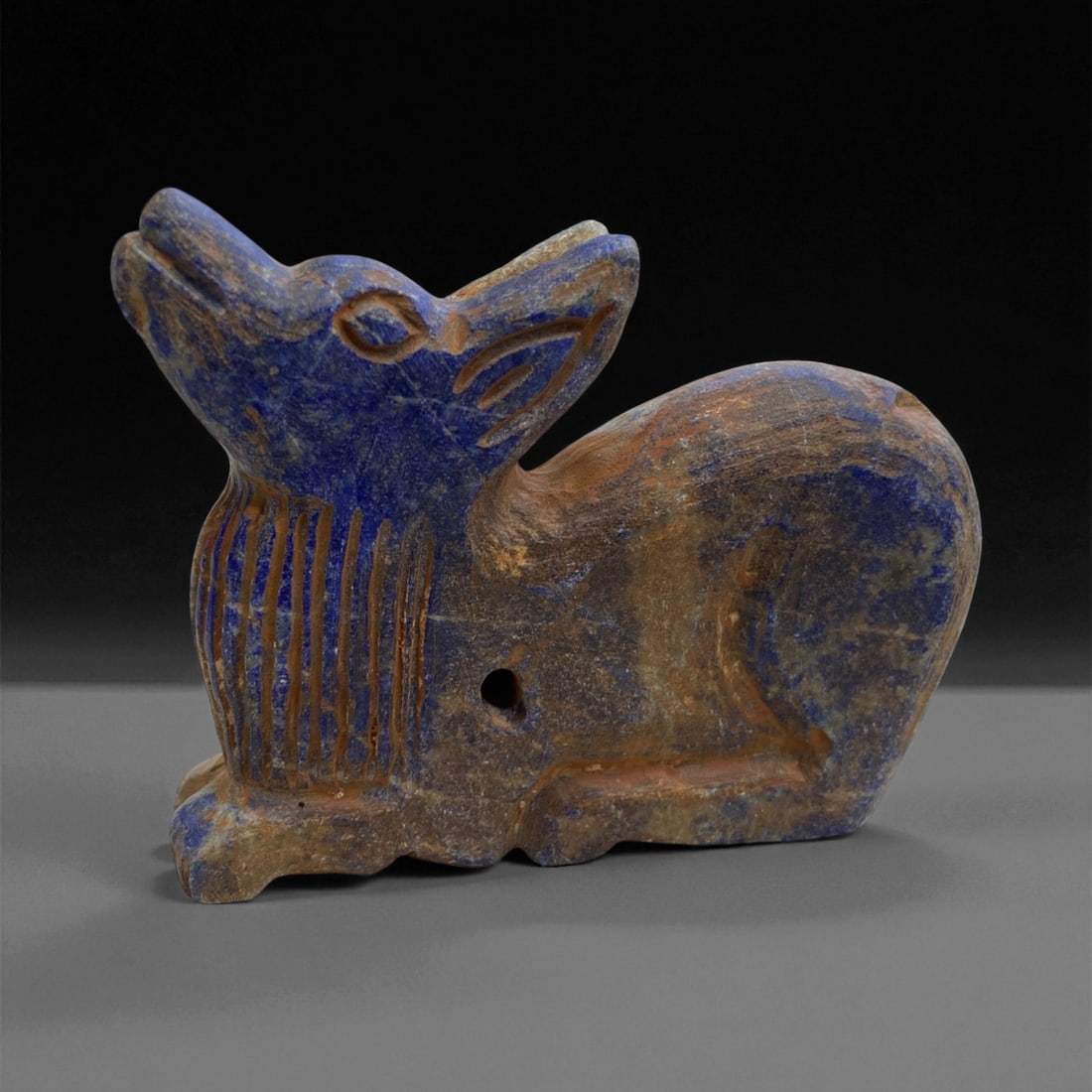 A Near Eastern Lapis Lazuli Zoomorphic Effigy, 2nd Millennium BCE, 138g, 28EE: Carved from lapis lazuli with deep blue areas and brown surface alterations, the effigy depicts a recumbent animal with the head raised and the snout projecting upward. Incised lines articulate the ma
