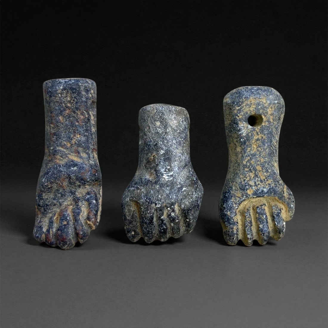 A Group of Three Egyptian Schist Fist Amulets, Late Period, 21 g, 35EE: Carved from dark grey schist with lighter surface mottling, each amulet is shaped as a clenched human fist with the thumb positioned along the side of the hand. The forms vary slightly in proportion,
