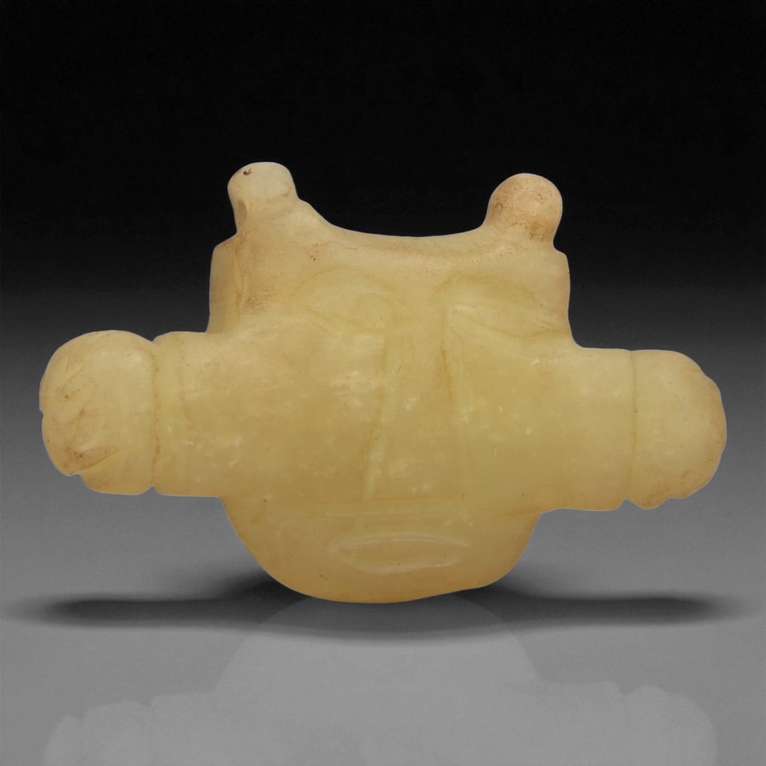 A Persian Jade Amulet, Medieval Period, 12th-14th Century CE, 15LE: Carved from a pale yellow jade with semi-translucent qualities, the amulet is shaped as a stylized human head with broad lateral projections. The facial features are indicated by shallow incised lines