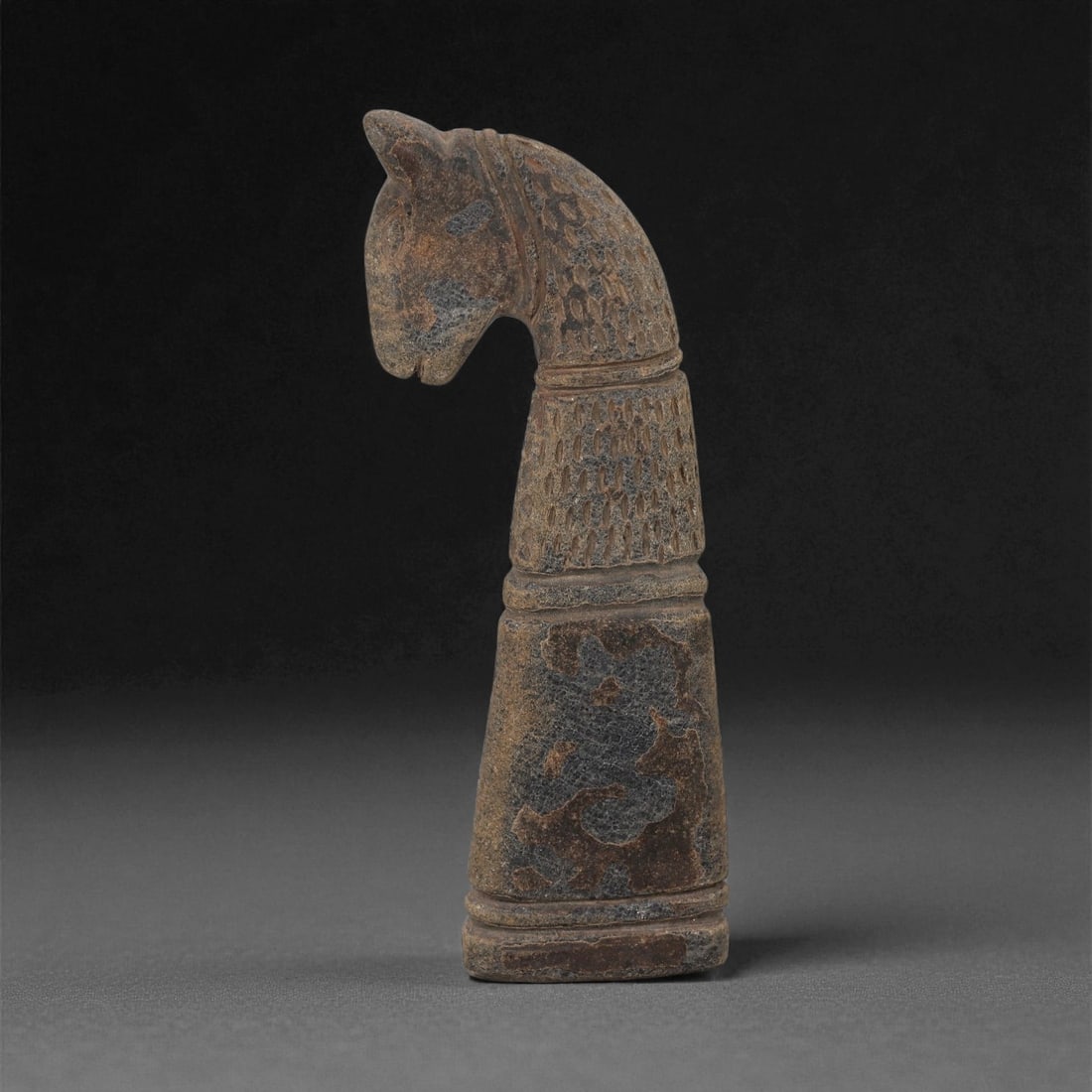 A Near Eastern Carved Stone Zoomorphic Effigy, Early 1st Millennium BCE, 38LE: Carved from a dark brown stone with lighter surface accretions, the effigy is shaped as an upright animal with an elongated, forward-curving neck and a narrow head with small rounded ears. The upper s