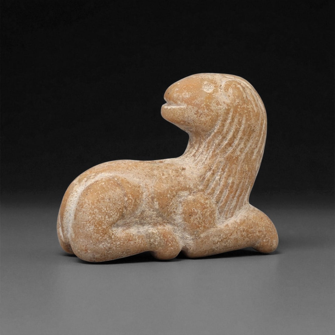 A Near Eastern Stone Recumbent Feline Amulet, 1st Millennium BCE, 12LE: Carved from a light brown stone with surface encrustations, the amulet depicts a recumbent feline with the forelegs folded beneath and the hindquarters tucked to one side. The head is raised and turne