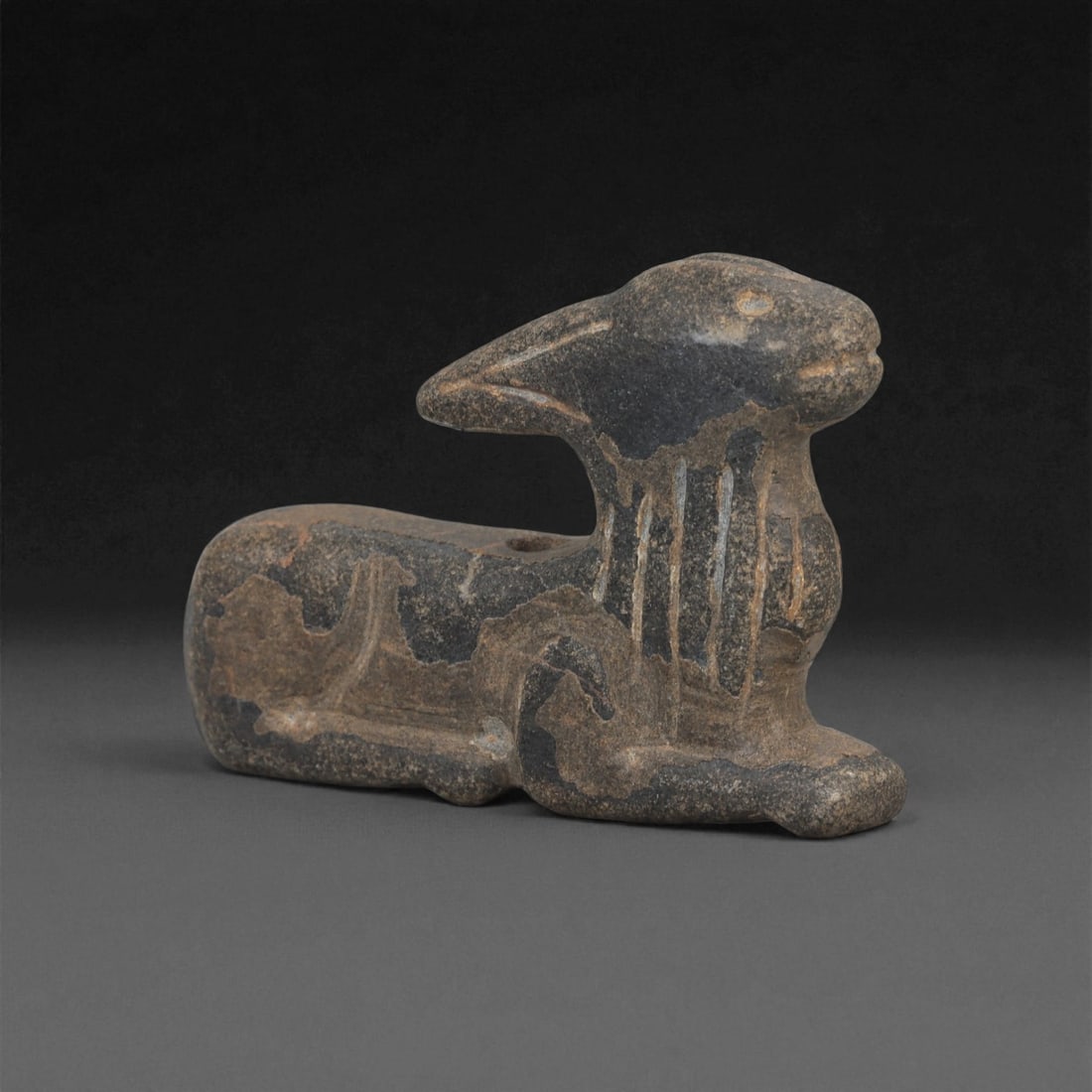 A Near Eastern Stone Zoomorphic Effigy, 3rd-2nd Millennium BCE, 90LE: Carved from a dark stone with lighter mottled surface patches, the object represents a recumbent quadruped with the forelegs extended and the hindquarters folded beneath the body. The head is elongate