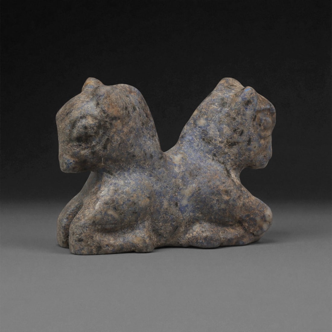 An Indo-Parthian Stone Double Zoomorphic Effigy, 1st-3rd Century CE, 55ZA: Carved from a dark stone with blue-grey surface mottling, the object depicts two conjoined quadrupeds shown back-to-back, each rendered with a rounded head, short muzzle, and simplified limbs tucked b