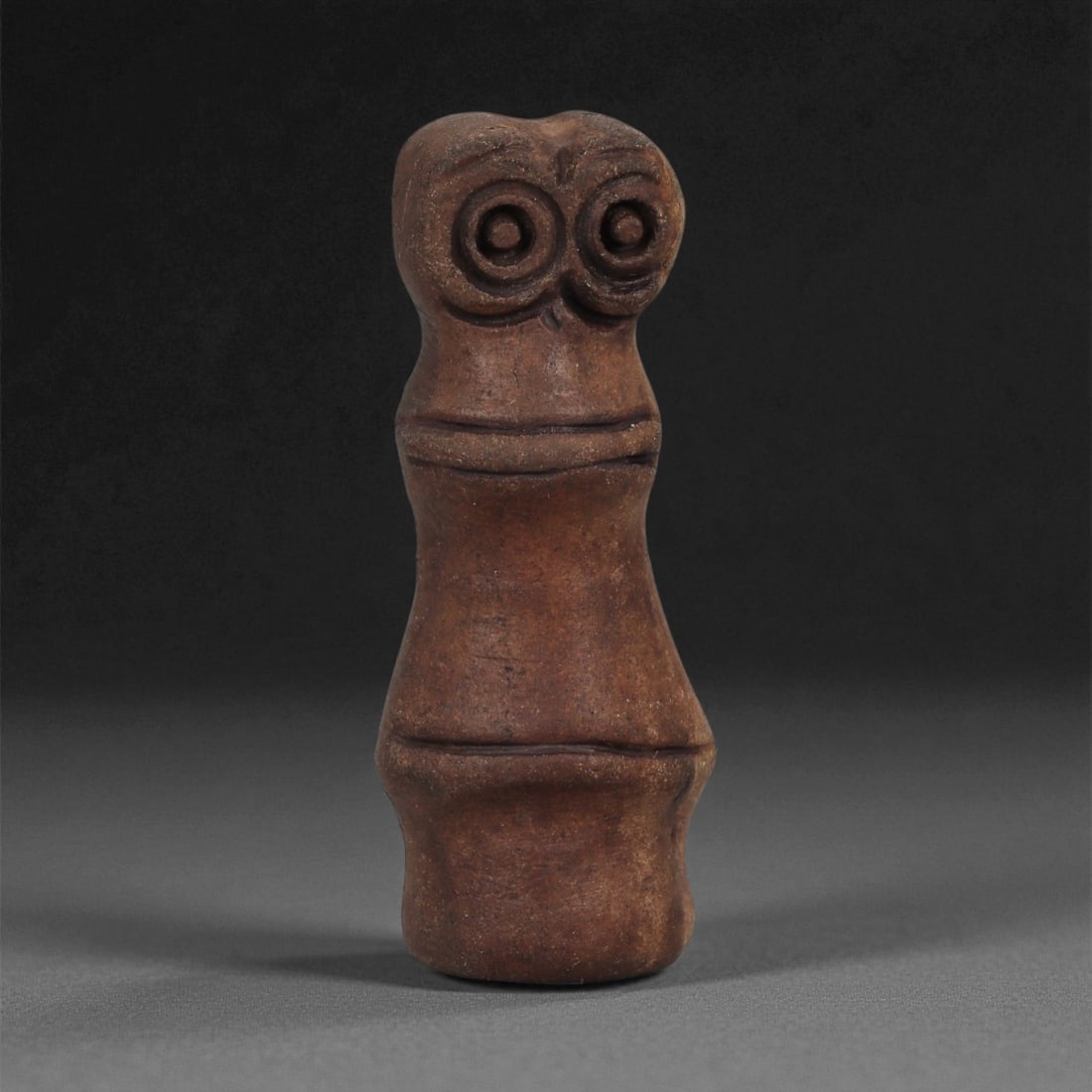 A Near Eastern Stone Eye Amulet, 3rd-2nd Millennium BCE, 86LE: Carved from a reddish-brown stone with a smooth, slightly burnished surface, the object is shaped as a tapering columnar form surmounted by a stylized head. The upper section bears two deeply recessed
