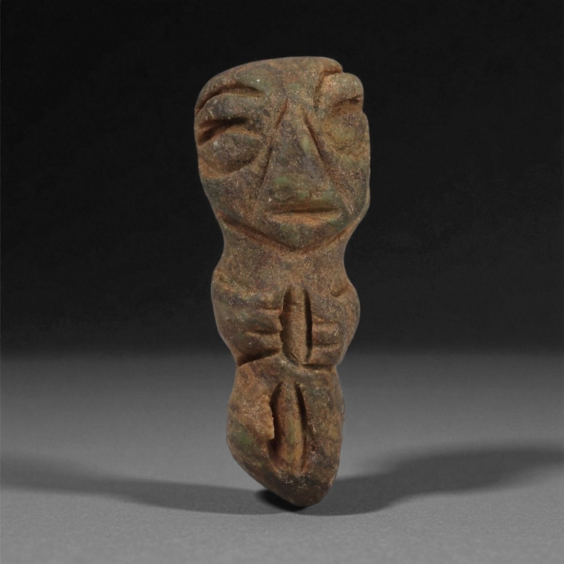 A Taino Carved Stone Idol, Late Pre-Columbian Period, Circa 1000-1500 CE, 77LF: Carved from a mottled brown-green stone with natural surface variations, the figure presents a tapering lower body incised with a vertical groove. The torso is defined by the arms brought forward, mee