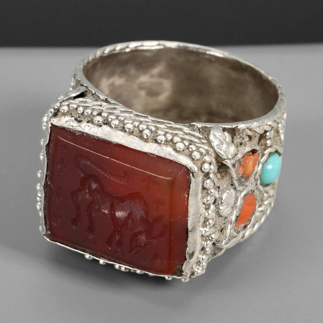 A Central Asian Silver Ring with Carnelian Intaglio and Coral-Turquoise Inlay, c. 19th-20th, 68XD: This impressive A silver ring captures the symbolic artistry of Central Asian jewelry from the late 19th to early 20th century. The rectangular carnelian intaglio at its center is engraved with a styl