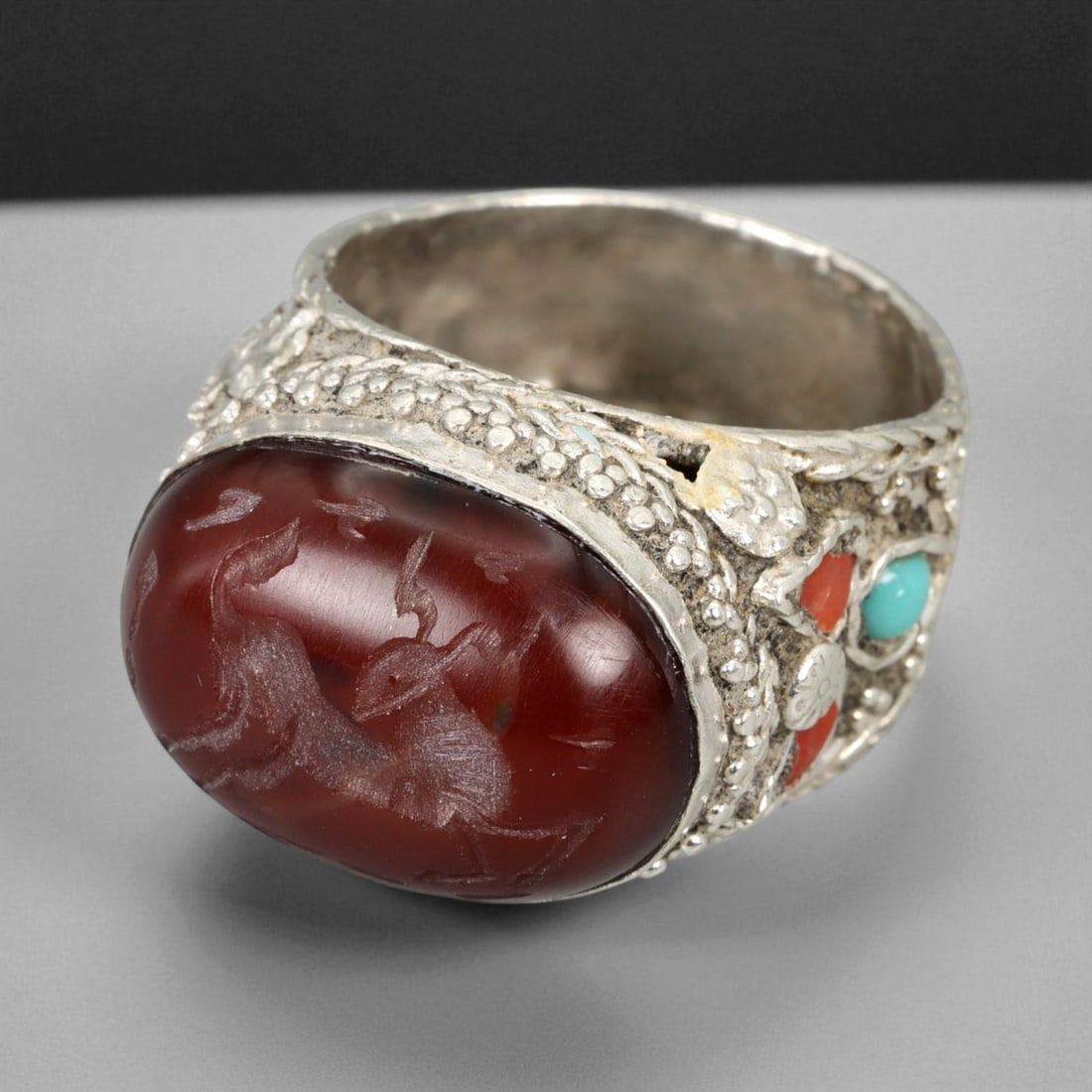 A Central Asian Silver Ring with Carnelian Intaglio and Turquoise Inlay, c. 19th-20th Century,, 16XD: This magnificent A silver ring embodies the regal aesthetic of Central Asian jewelry from the late 19th to early 20th century. The centerpiece, a large oval carnelian intaglio, is finely engraved with