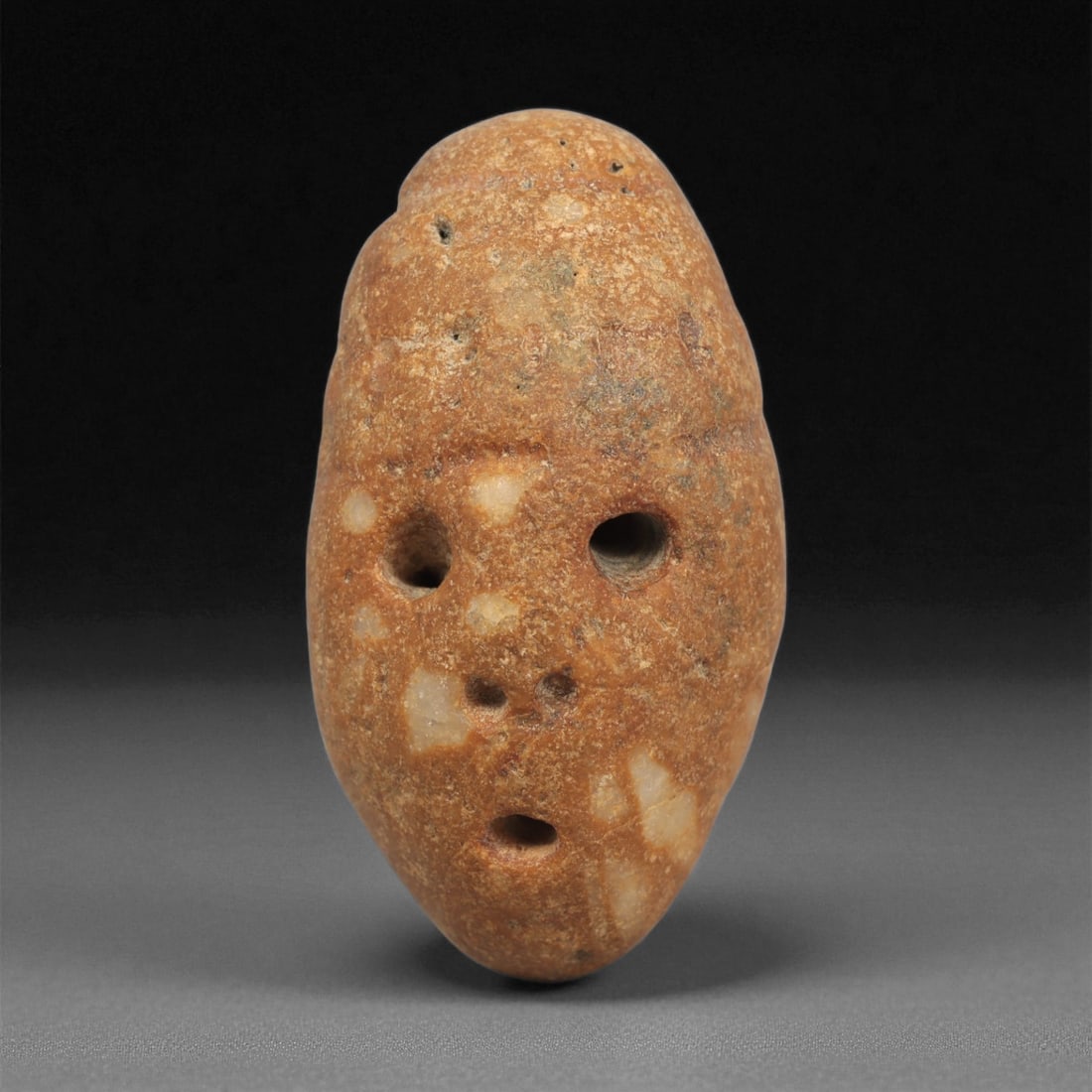 A Mediterranean Stone Maskette, 12th-17th Century, 94LC: Carved from a mottled orange-brown stone with lighter quartz inclusions, the maskette is shaped with an elongated oval contour tapering toward a narrow chin. The facial features are defined by drilled