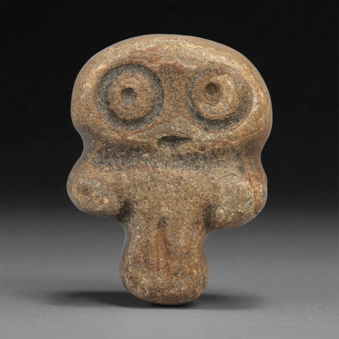 A Near Eastern Stone Anthropomorphic Figurine, 4th-3rd Millennium BCE, 6LD: Carved from a brown stone with mottled surface coloration, the figurine is modeled with a broad rounded head, large circular recessed eyes with central drilling, and a small incised mouth below. The s