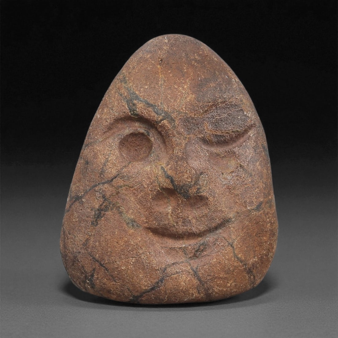 A Mediterranean Stone Maskette, 12th-17th Century, 72LC: Carved from a reddish-brown stone with dark veining, the maskette is shaped with a broad, triangular outline tapering gently toward the base. The features are rendered in low relief, including one rec