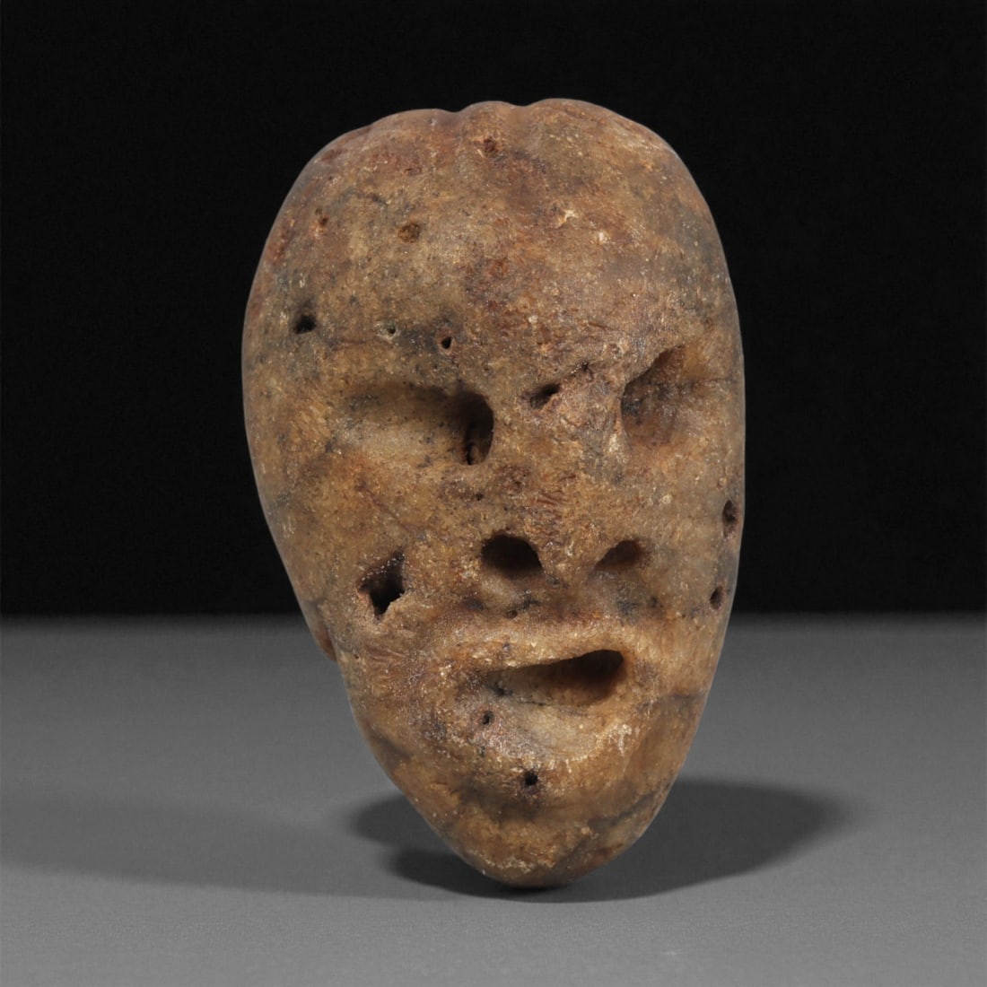 A Mediterranean Stone Maskette, 12th-17th Century, 5LF: Carved from a light brown stone with scattered natural cavities and surface pitting, the maskette is shaped with an elongated, tapering form. The facial features are minimally defined, with recessed o