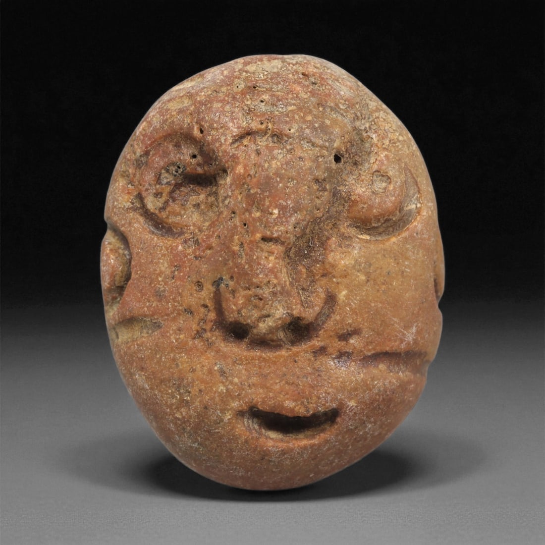 A Mediterranean Stone Maskette, 12th-17th Century, 24LC: Carved from a reddish-brown stone with scattered surface pitting and natural inclusions, the maskette is shaped with an oval contour tapering slightly toward the chin. The facial features are rendered