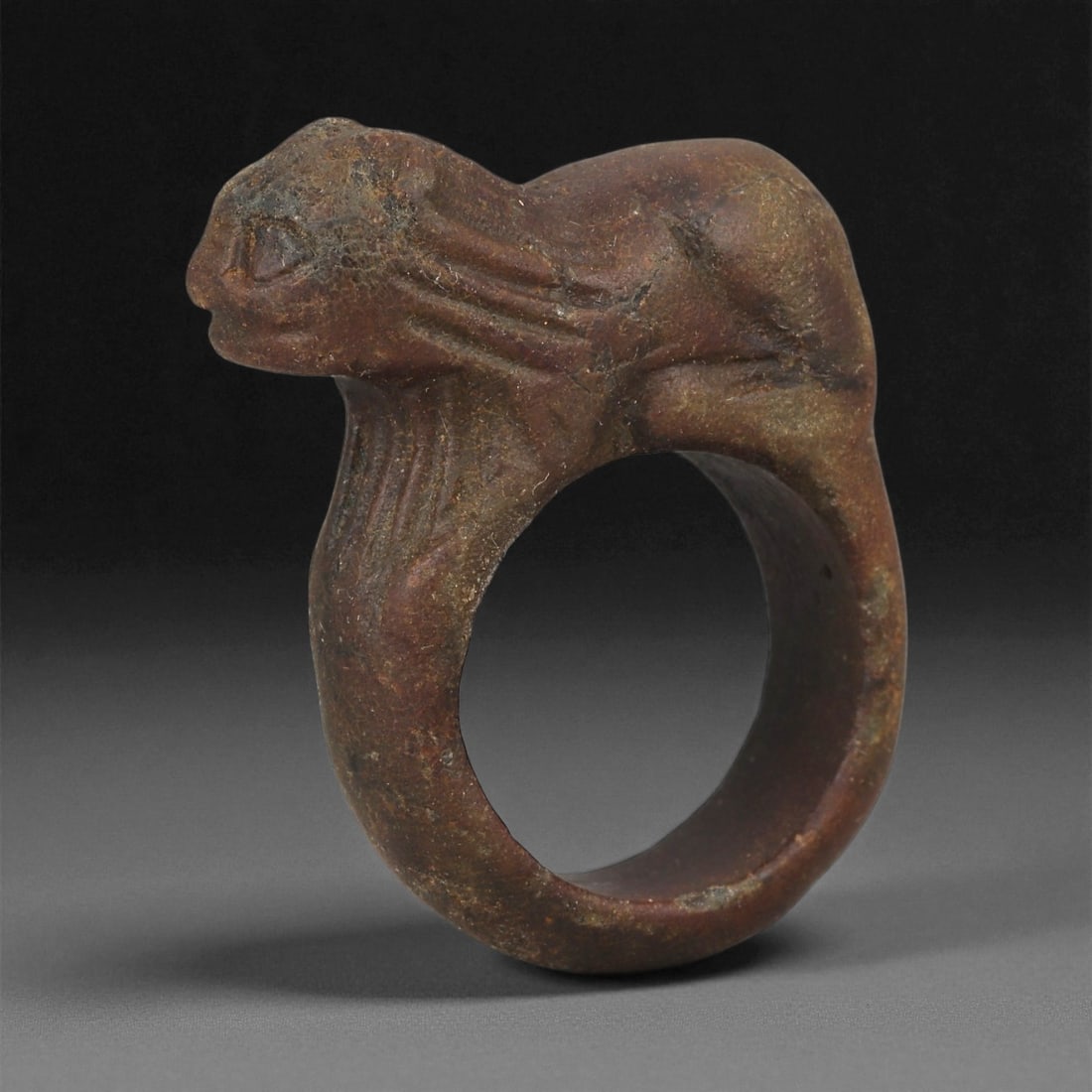 An Indo-Parthian Stone Zoomorphic Ring, 1st-2nd Century CE, 40LD: Carved from brown stone with lighter surface inclusions, the ring is formed with a circular hoop surmounted by a recumbent feline rendered in low relief. The head is shaped with a short snout and rece