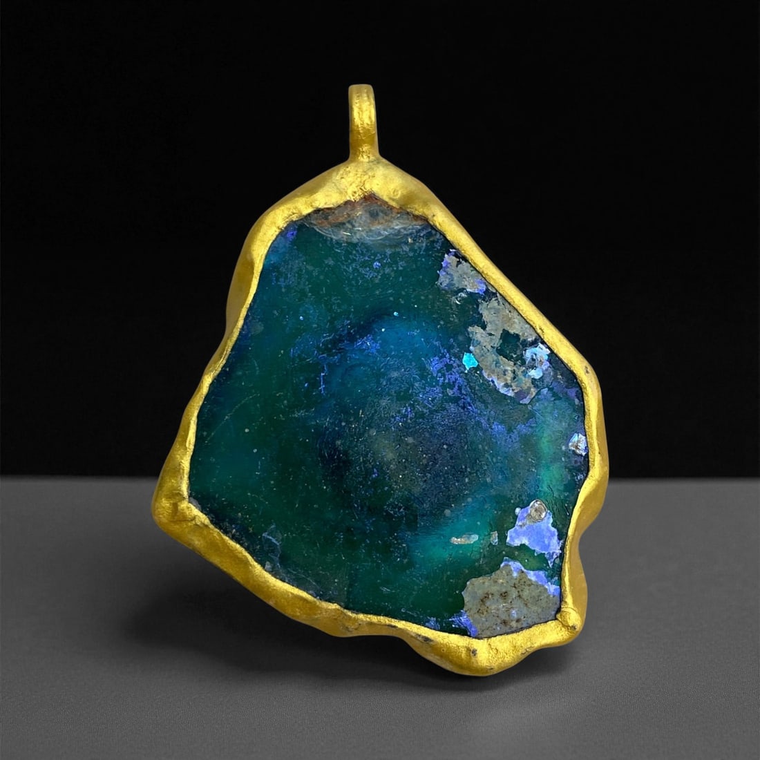 A Roman Gold-Mounted Glass Pendant, 1st-3rd Century CE, 35g, 9EE: Formed from a fragment of blue-green Roman glass with areas of iridescence and weathering, the pendant is set within a custom-fitted gold frame that follows the irregular outline of the shard. The mou