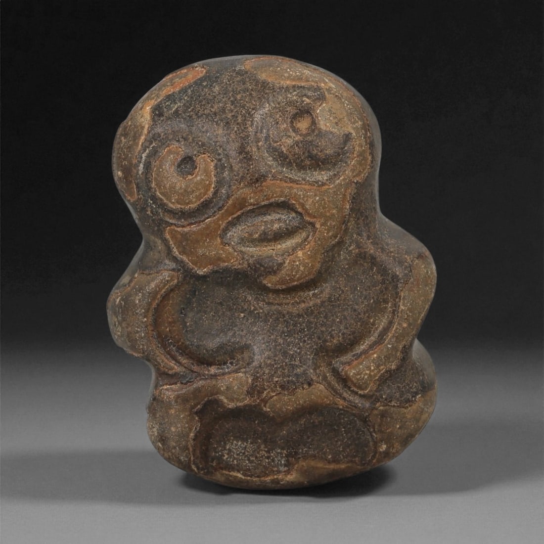 A Near Eastern Stone Anthropomorphic Figurine, 3rd-2nd Millennium BCE, 2LB: Carved from a dark brown stone with lighter surface mottling, the object depicts a highly stylized anthropomorphic figure with a rounded head and prominent circular eyes defined by concentric grooves.