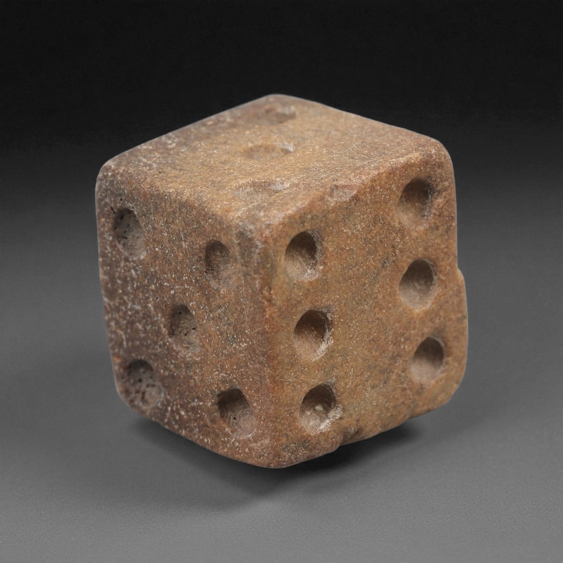 A Roman Bone Gaming Die, 1st-3rd Century CE, 55LD: Carved from light brown bone with a porous, naturally textured surface, the cubic die bears circular recessed pips on each face arranged in standard numerical configurations. The edges are slightly ro