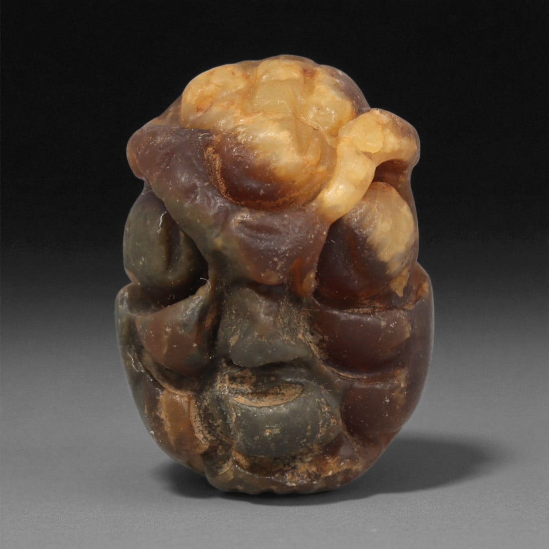 A Mediterranean Stone Maskette, 12th-17th Century, 20LC: Carved from a variegated stone with translucent amber-brown and opaque grey tonal zones, the maskette has a compact, rounded form. The facial features are modeled in shallow relief, including recessed