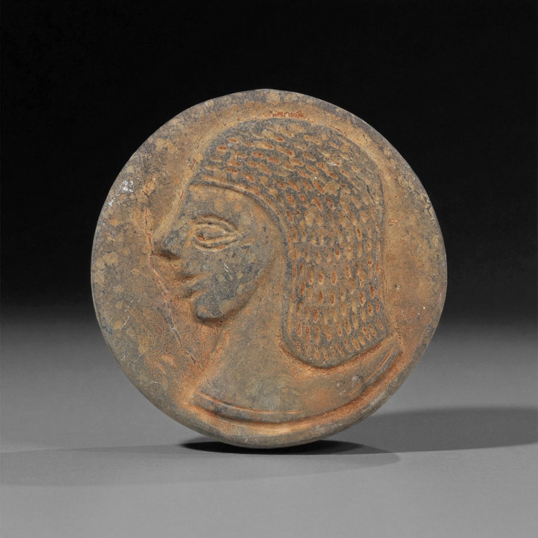 An Egyptian Stone Plaque with Male Profile, New Kingdom, 1550-1070 BCE, 85ZA: Carved from a mottled brown stone with surface encrustations, the circular plaque bears a profile head facing left. The figure has a prominent eye rendered with a recessed outline and a straight brow.