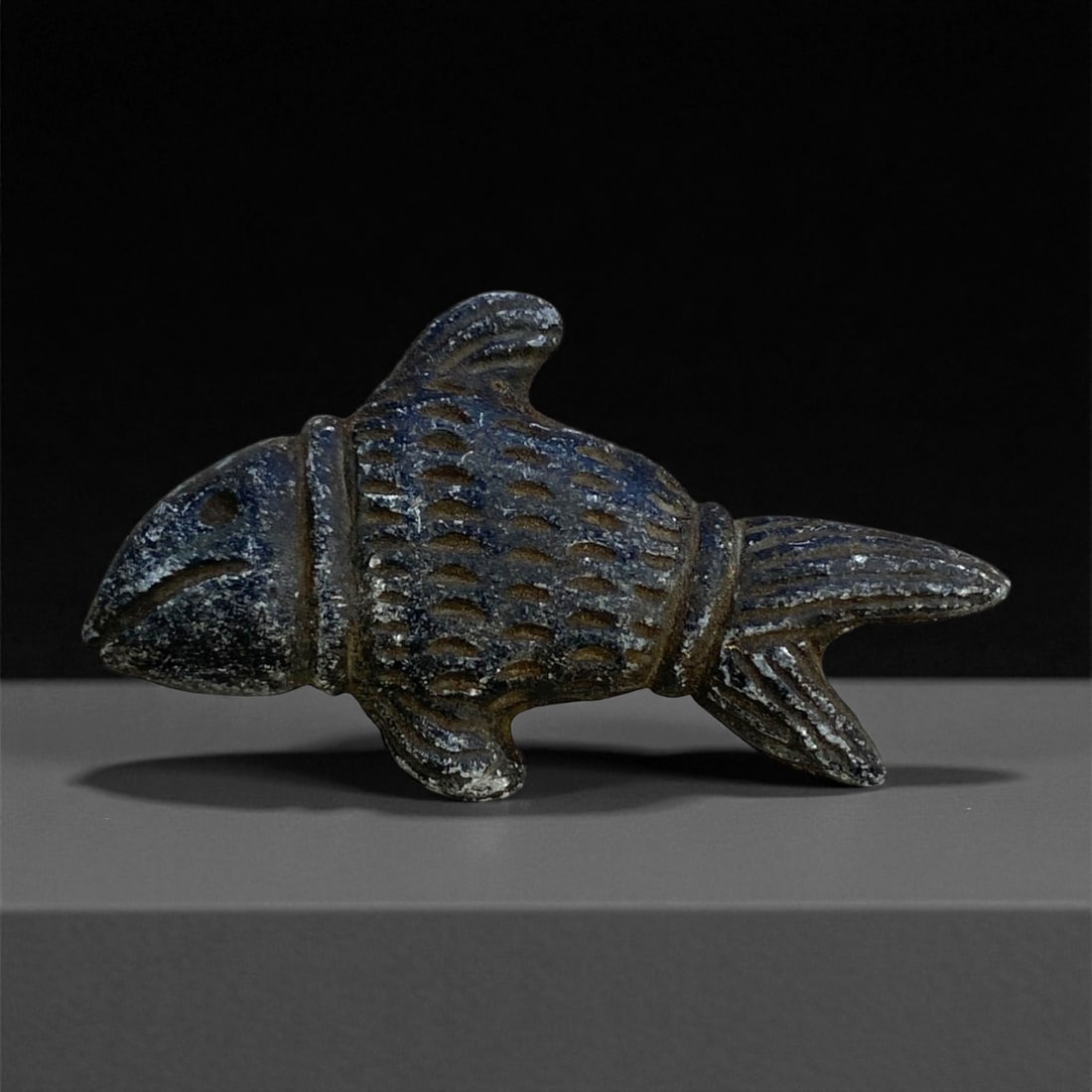 A Near Eastern Stone Fish Effigy, 1st Millennium BCE, 26g, 16EE: Carved from dark stone with a fine granular surface, the effigy is shaped as a stylized fish with a rounded head, incised eye, and a linear groove marking the mouth. The body is defined by a series of
