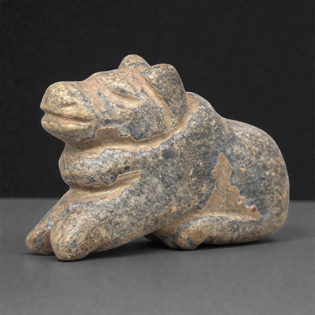 A Near Eastern Stone Zoomorphic Effigy, 3rd-2nd Millennium BCE, 72LF: Carved from a dark grey stone with lighter brown surface exposures, the figure depicts a recumbent quadruped with a compact body and short, rounded muzzle. Incised grooves delineate the head, limbs, a