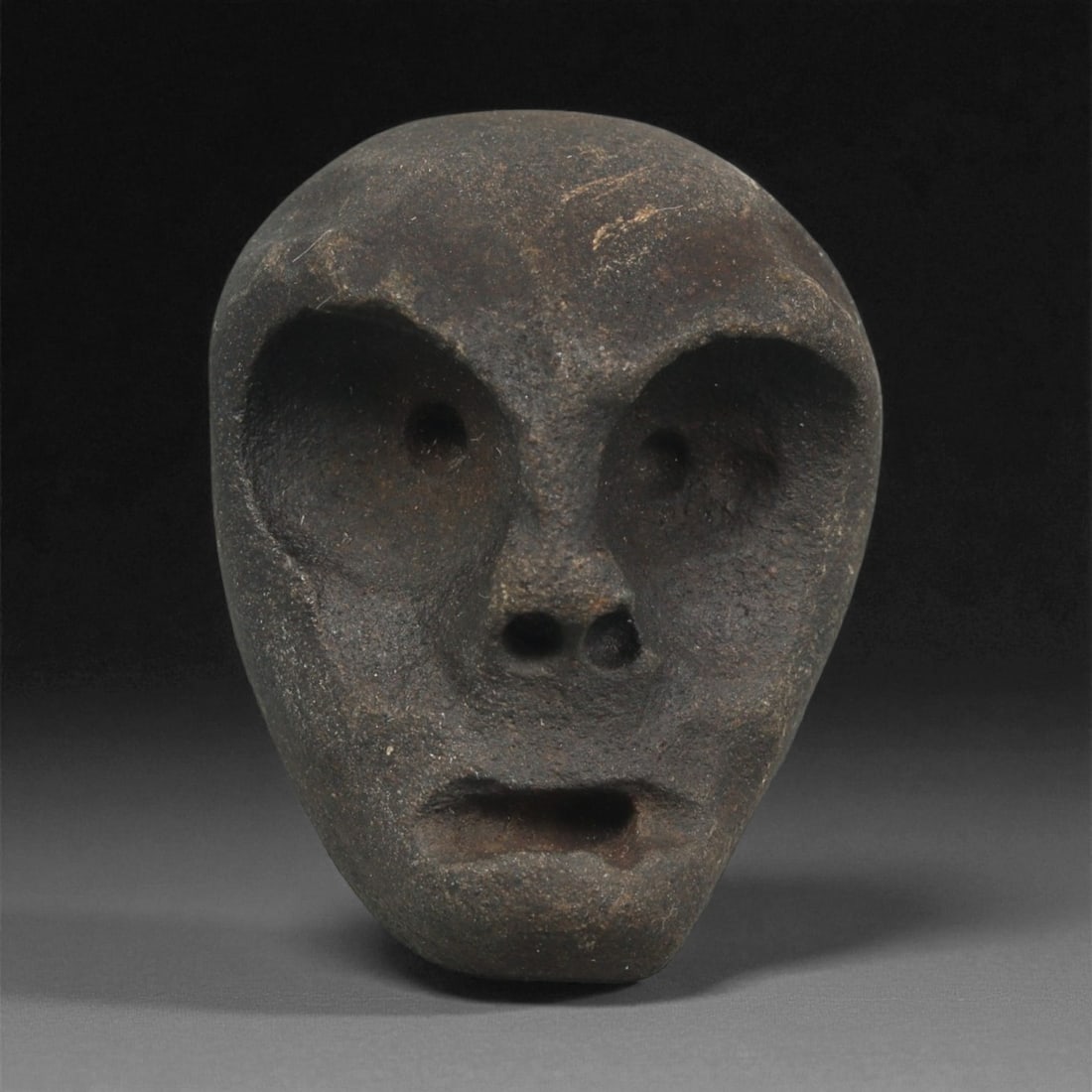 A Mediterranean Stone Maskette, 12th-17th Century, 13ZB: Carved from a dark brown stone with a dense, fine-grained surface, the maskette displays an elongated oval form tapering slightly at the chin. The facial features are rendered in deep relief, with lar