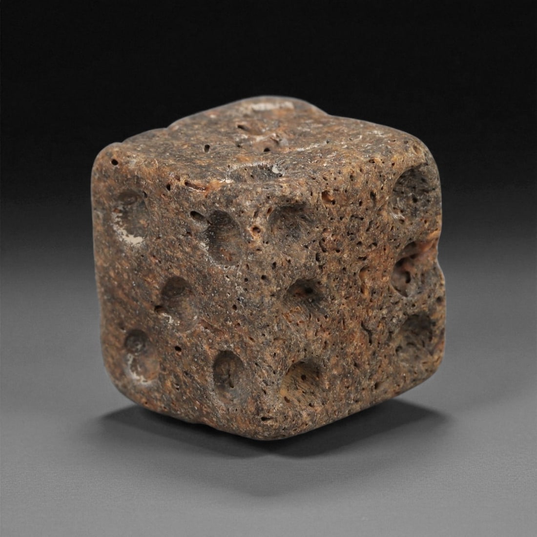 A Roman Bone Gaming Die, 1st-3rd Century CE, 63ZB: Carved from dense bone, the object is shaped as a small cube with slightly rounded edges. Each face bears a series of drilled circular pips arranged in standard numerical configurations. The surfaces 