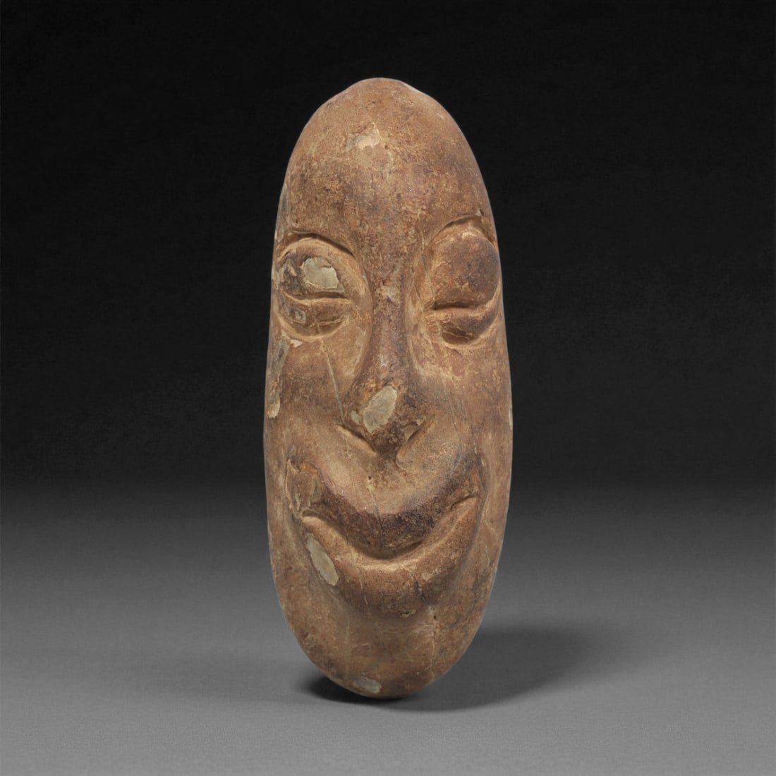 A Mediterranean Stone Maskette, 12th-17th Century, 11LF: Carved from a light brown stone with scattered pale surface inclusions, the maskette is shaped with an elongated oval outline and a smoothly rounded top. The facial features are modeled in shallow rel