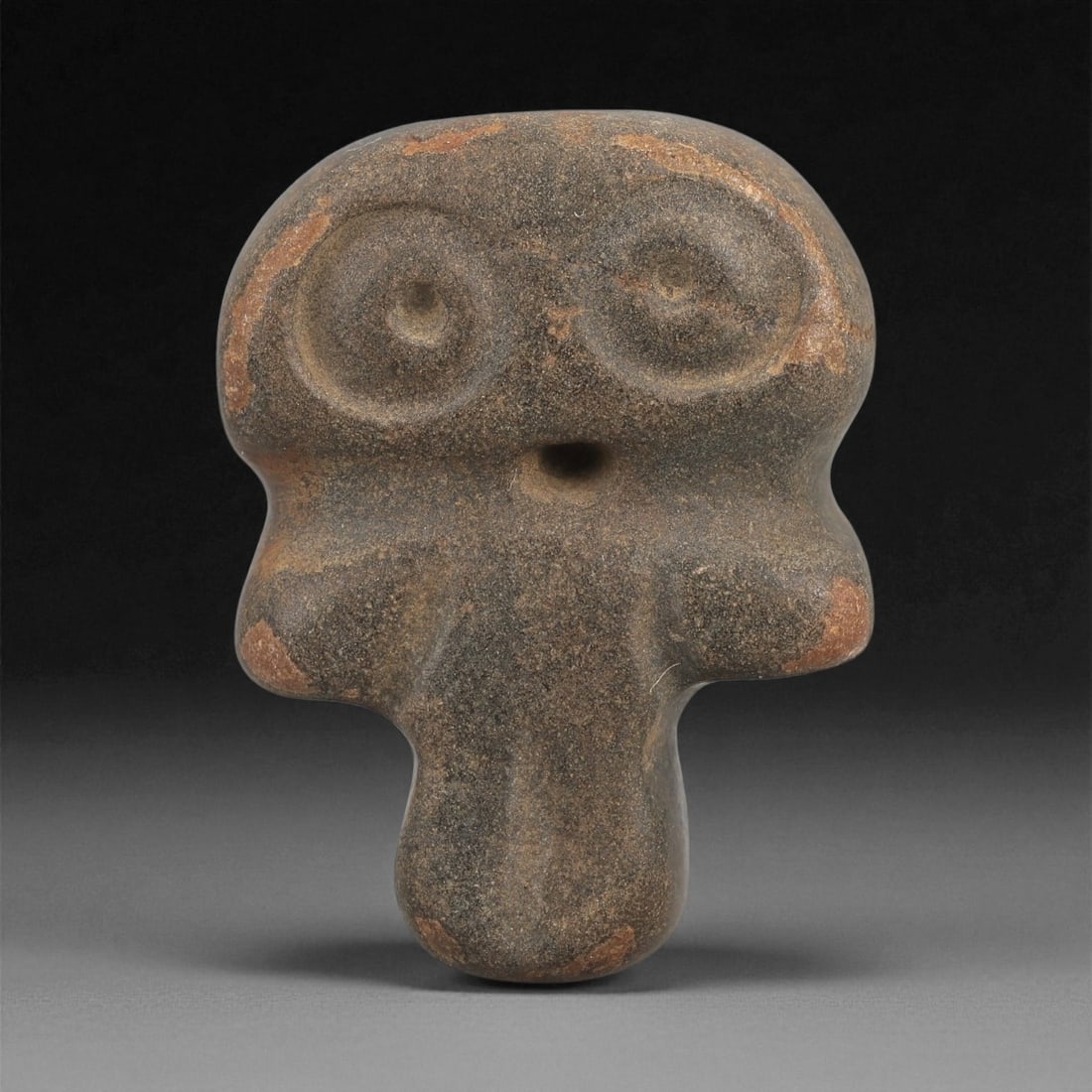A Near Eastern Stone Anthropomorphic Figurine, 4th-3rd Millennium BCE, 56LD: Carved from a dark brown stone with lighter surface abrasions, the figurine is modeled with a broad rounded head bearing two large circular recessed eyes and a small drilled mouth opening. The shoulde
