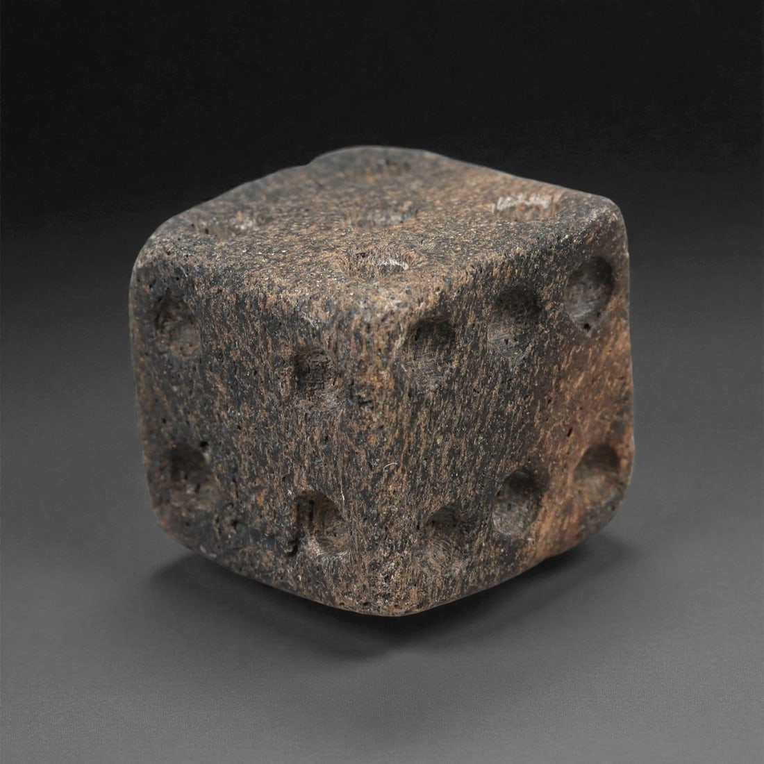 A Roman Bone Gaming Die, 1st-3rd Century CE, 86LD: Carved from light brown bone with a porous, naturally textured surface, the cubic die bears circular recessed pips on each face arranged in standard numerical configurations. The edges are slightly ro