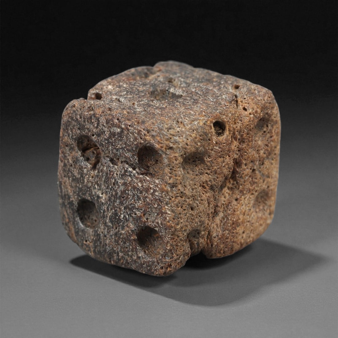 A Roman Bone Gaming Die, 1st-3rd Century CE, 30LD: Carved from light brown bone with a porous, naturally textured surface, the cubic die bears circular recessed pips on each face arranged in standard numerical configurations. The edges are slightly ro