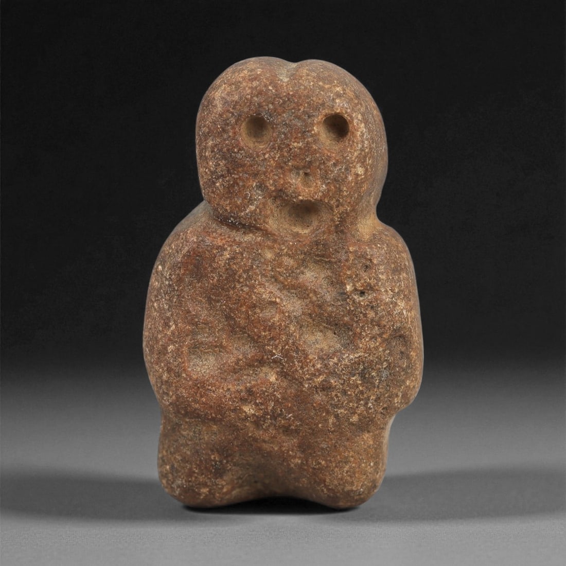 A Near Eastern Stone Anthropomorphic Figurine, 4th-3rd Millennium BCE, 24LD: Carved from a reddish-brown stone with surface weathering, the figurine is shaped with a rounded head, drilled circular eyes, and a small recessed mouth. The shoulders are slightly indicated, and the 
