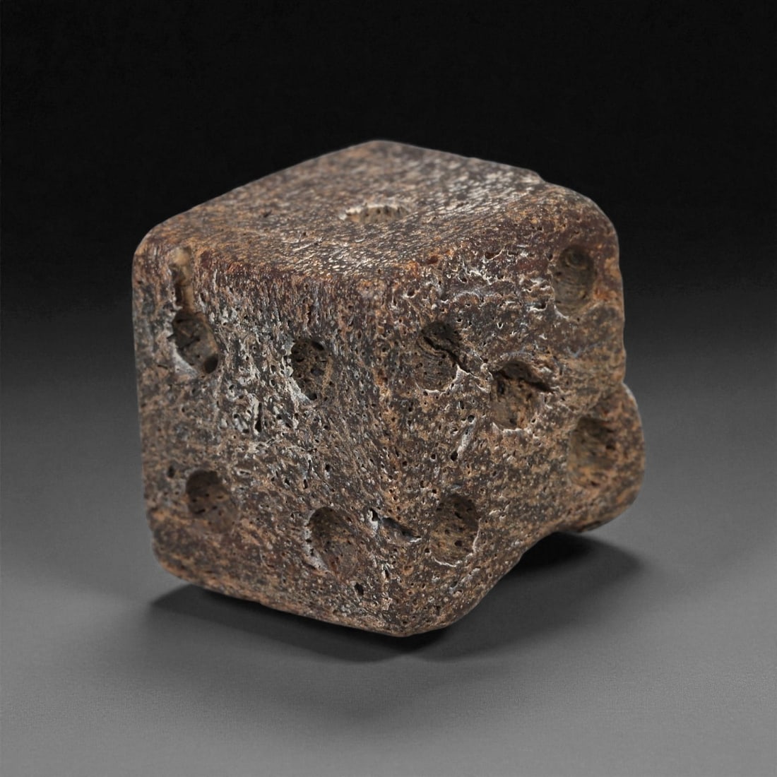 A Roman Bone Gaming Die, 1st-3rd Century CE, 53ZA: Carved from dense bone, the object is shaped as a small cube with slightly rounded edges. Each face bears a series of drilled circular pips arranged in standard numerical configurations. The surfaces 