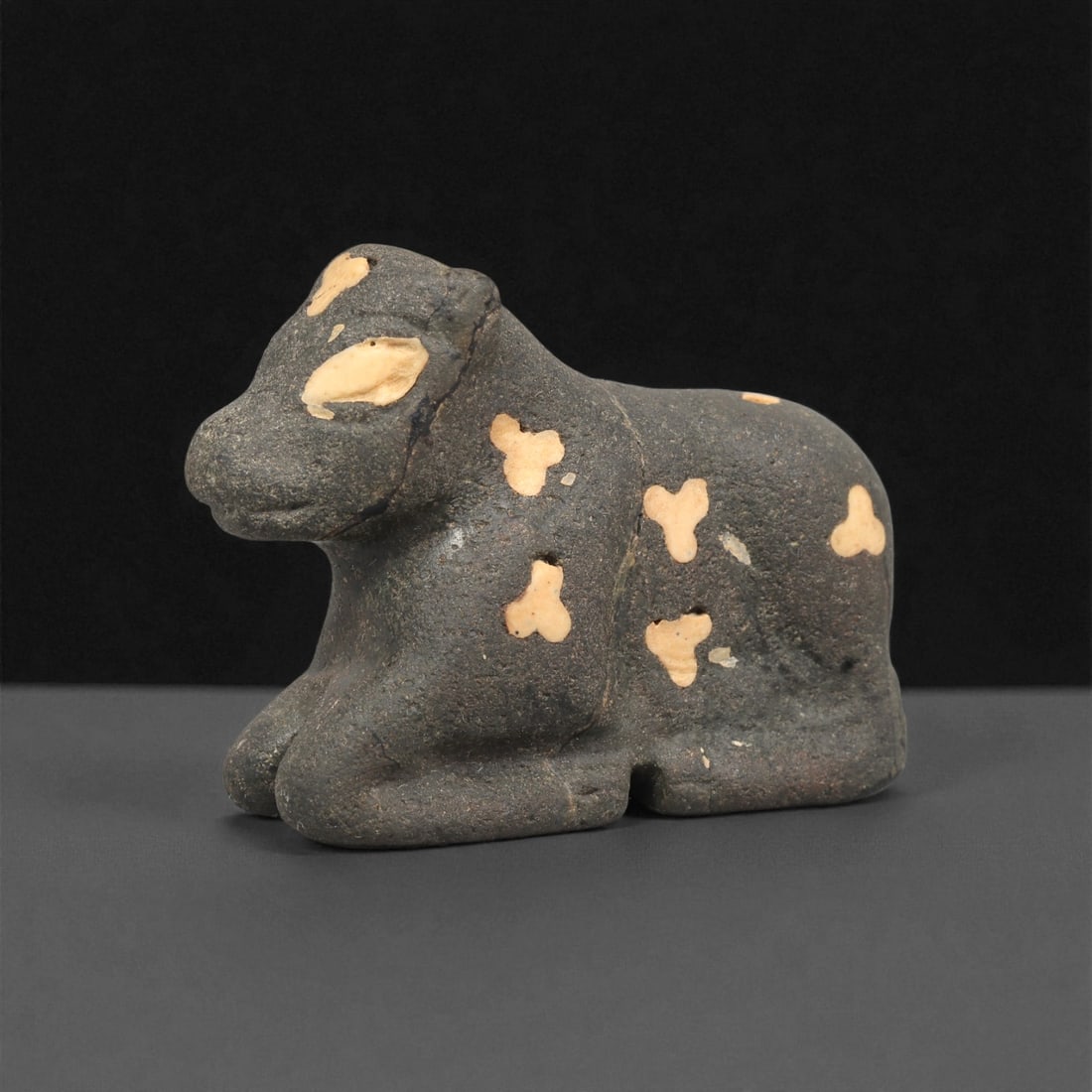 A Near Eastern Stone Bovid Effigy, 3rd-2nd Millennium BCE, 26ZA: Carved from a dark stone with lighter inclusions set into the surface, the object represents a recumbent bovid with its forelegs tucked beneath the body. The head is rounded with a short muzzle, and t