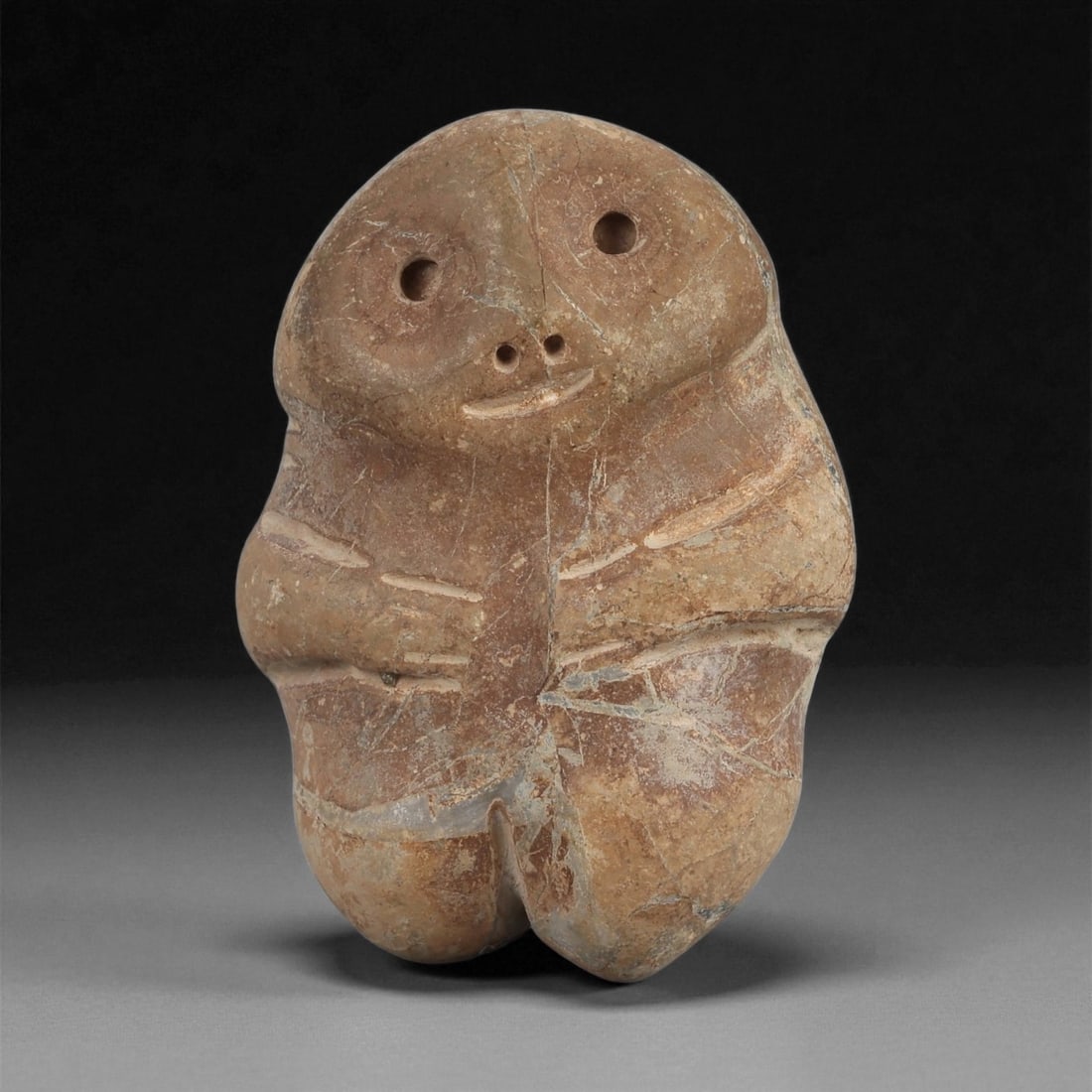 A Near Eastern Stone Anthropomorphic Figurine, 4th-3rd Millennium BCE, 9ZB: Carved from a light brown stone with surface veining, the figure is rendered with a rounded head and a compact body. The face displays two drilled circular eyes and a pair of small perforations sugges