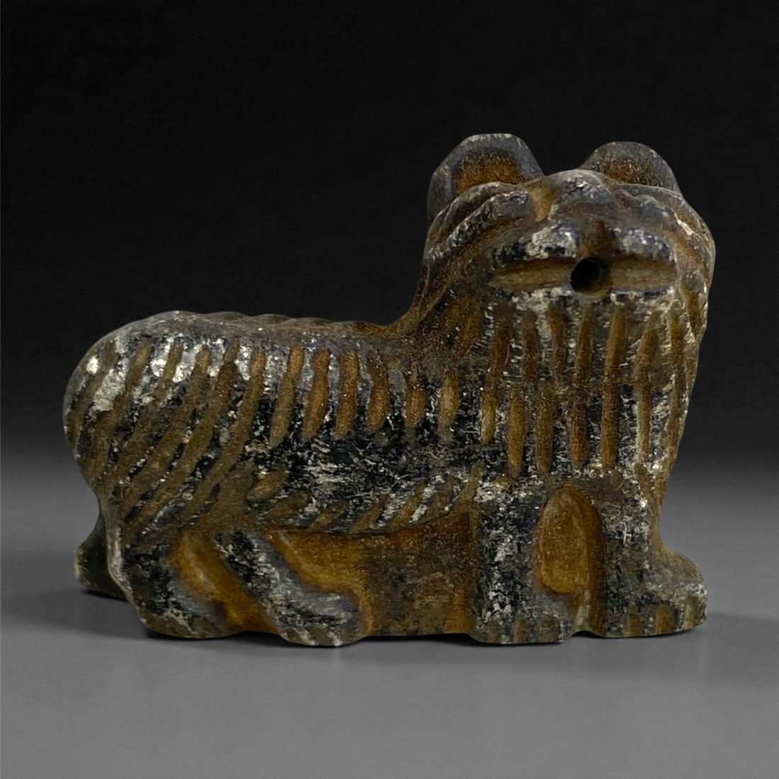 A Near Eastern Stone Zoomorphic Effigy, 2nd-1st Millennium BCE, 22g, 25EE: Carved from a dark stone with lighter veining, the effigy is shaped as a quadruped with a compact body and a short upturned head. Deep parallel incisions articulate the mane, flanks, and legs, while a