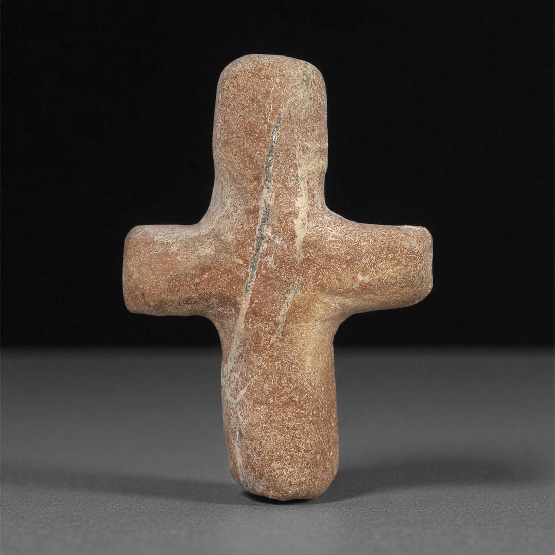 An Early Christian Stone Cross Pendant, 5th-7th Century CE, 59ZA: Carved from a brown stone with natural veining, the object is shaped as a compact cross with rounded terminals. The arms and shaft are smoothly contoured, with no additional carved ornamentation. The 
