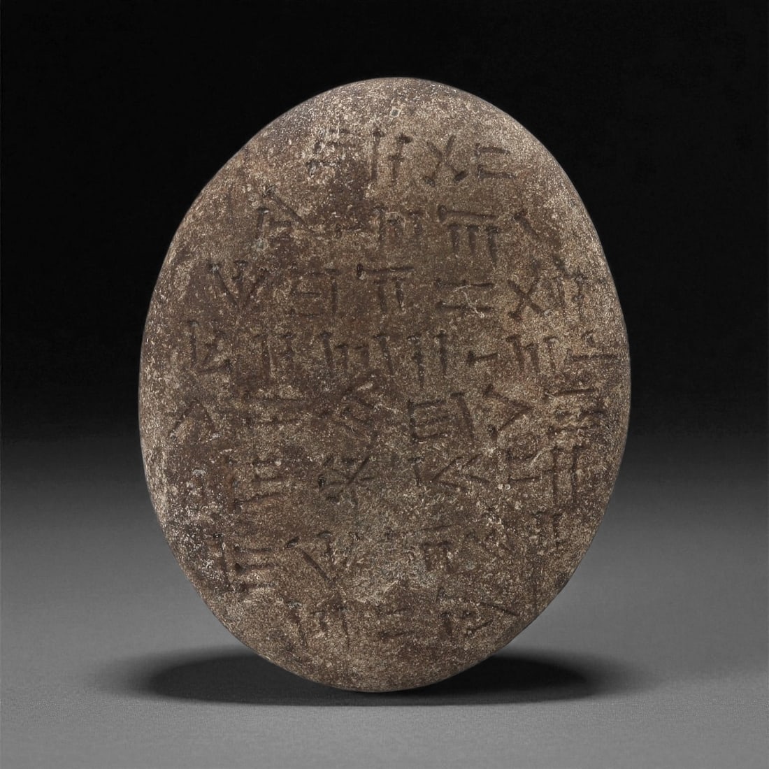 A Sumerian Inscribed Stone Plaque, Early-Mid 2nd Millennium BCE, 36LE: Carved from a dark brown stone with a slightly flattened ovoid profile, the surface bears multiple lines of incised cuneiform signs arranged in loose horizontal groupings. The inscription consists of 