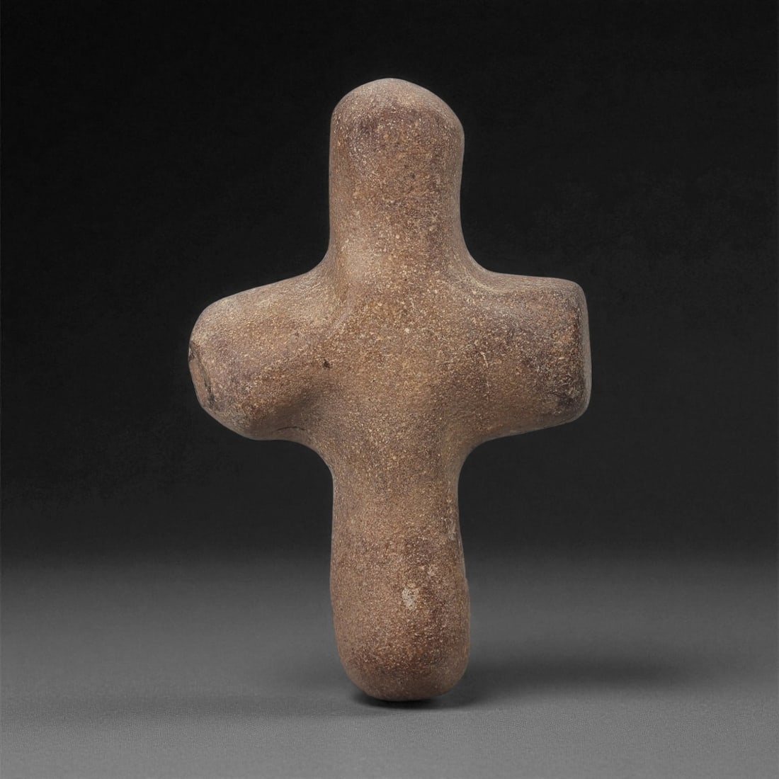 A Byzantine Stone Cross, 5th-7th Century CE, 76LB: Carved from a dark brown stone with natural surface veining, the object is shaped as a cross with rounded terminals and a tapering lower section. The front preserves shallow linear carving forming a c
