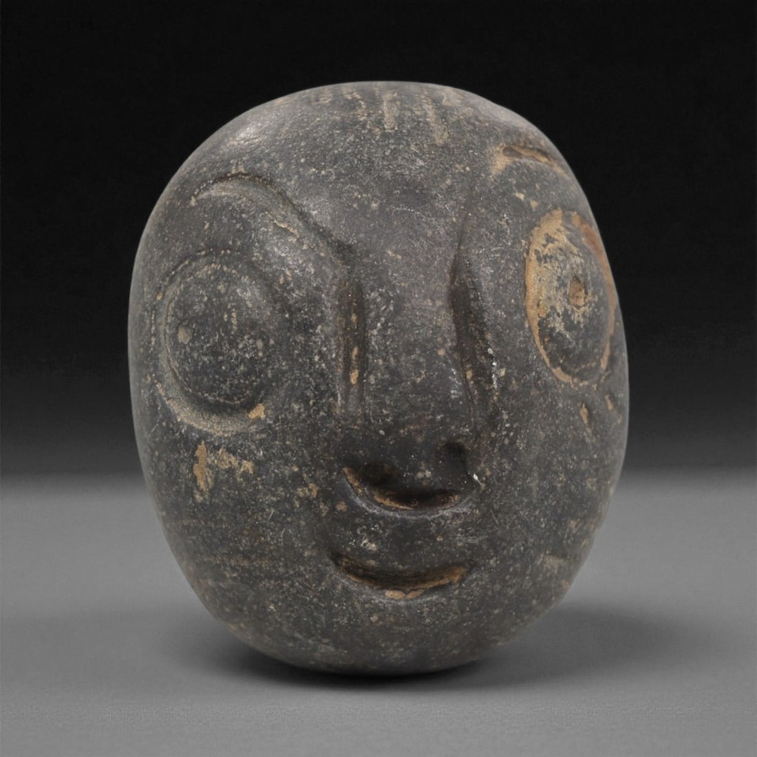 A Mediterranean Stone Maskette, 12th-17th Century, 54LC: Carved from a dark grey stone with lighter inclusions and minor surface abrasions, the maskette is shaped with a rounded, globular outline. The facial features are rendered in shallow relief, includin