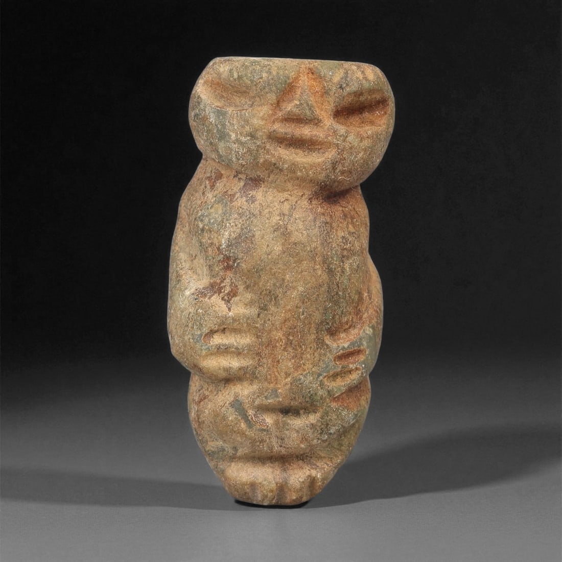A Taino Carved Stone Idol, Late Ceramic Age, circa 1200-1500 CE, 80LE: Carved from mottled brown stone with areas of greenish surface accretion, the figure is modeled standing with a rounded lower body and short legs. The head is broad and flattened at the top, featuring