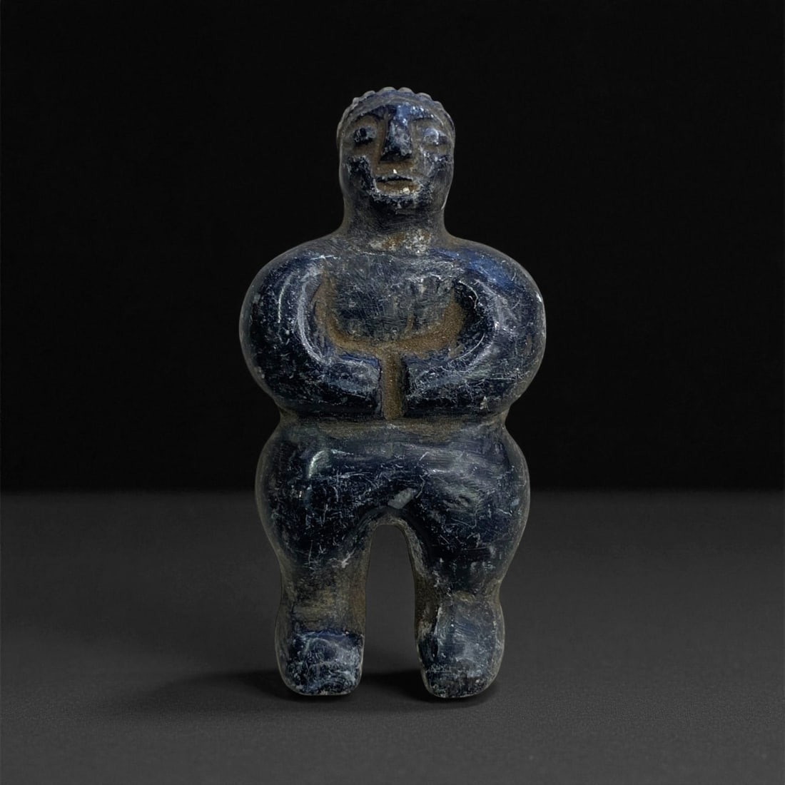 A Near Eastern Stone Anthropomorphic Idol, 2nd-1st Millennium BCE, 27g, 17EE: Carved from dark stone with a smooth, slightly glossy surface, the figure is modeled with a rounded head, prominent nose, and recessed eyes. The arms are bent forward and joined at the hands over the 