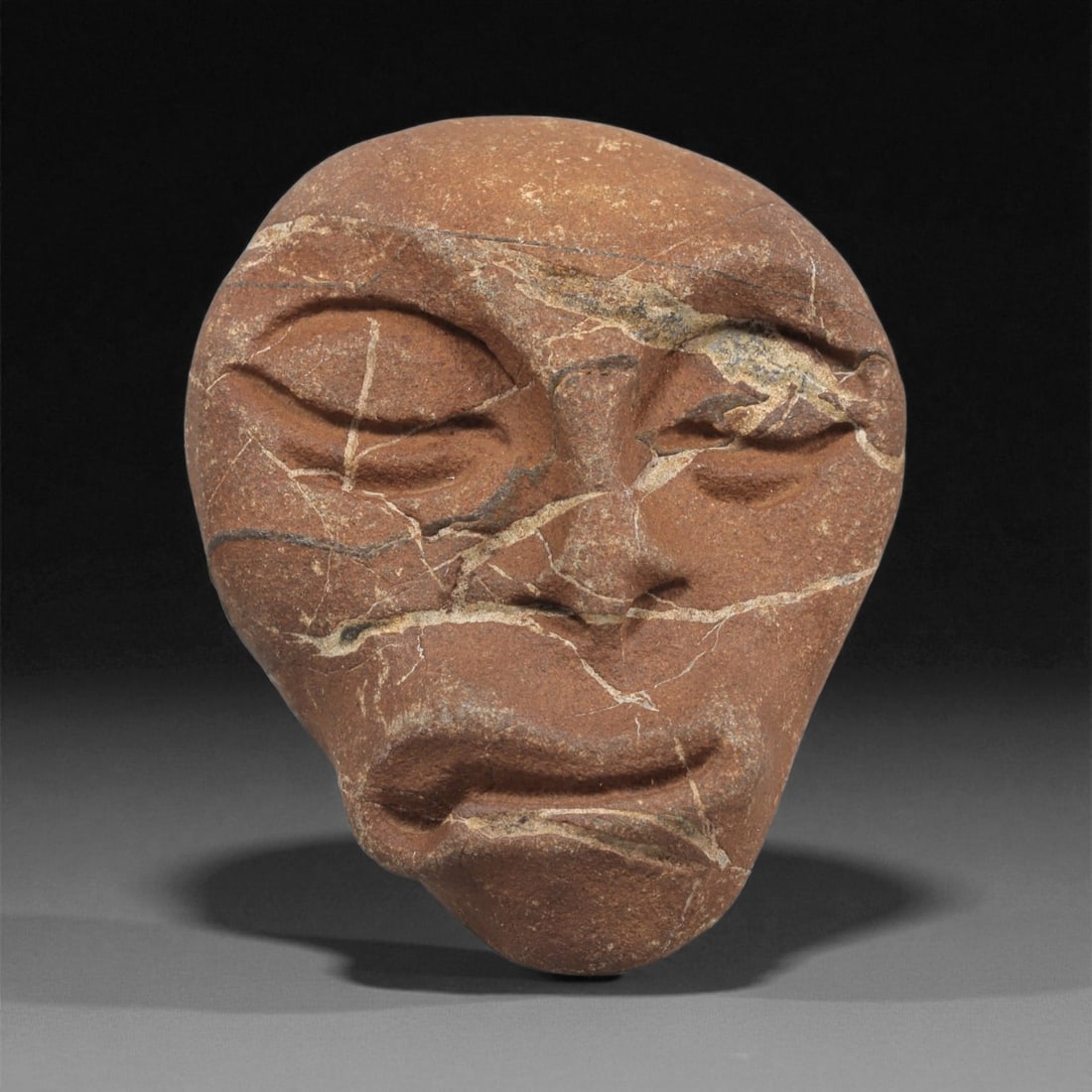 A Mediterranean Stone Maskette, 12th-17th Century, 50ZA: Carved from a reddish-brown stone with extensive natural veining, the maskette is shaped with a broad upper section narrowing toward the chin. The facial features are rendered in shallow relief, inclu