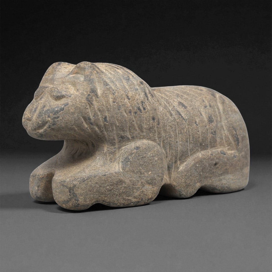 A Near Eastern Stone Feline Effigy, 3rd-2nd Millennium BCE, 70LF: Carved from a brown-grey stone with darker inclusions, the figure depicts a recumbent feline with a rounded head and short projecting snout. Incised lines articulate the mane, flank, and limbs, while 