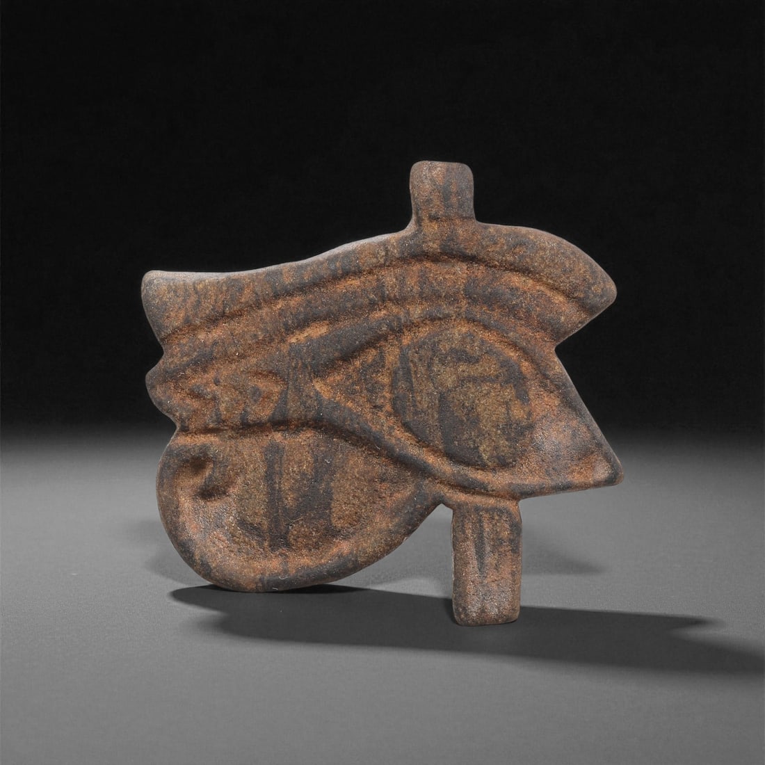 An Egyptian Stone Wedjat Eye, Late Period, 664-332 BCE, 66LF: Carved from a dark brown stone with lighter surface mottling, the object is formed as a stylized wedjat eye with the characteristic extended cosmetic lines above and below the eye and a vertical eleme