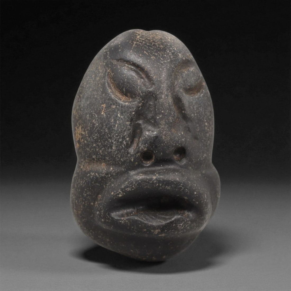 A Mediterranean Stone Maskette, 12th-17th Century, 66LC: Carved from a dark grey stone with fine granular inclusions and scattered surface abrasions, the maskette has an elongated oval outline with a slightly bulbous lower section. The facial features are r